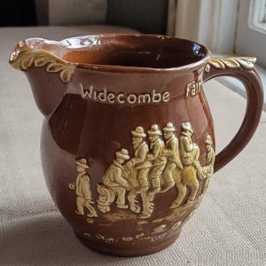 May include: A brown ceramic pitcher with a handle and a scene of people riding a horse. The pitcher has the words "Widecombe Fair" on the side.