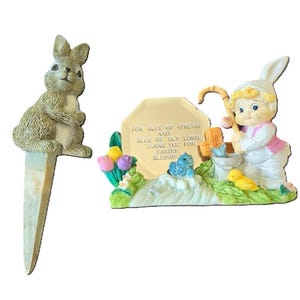 May include: A set of two Easter figurines. One is a brown rabbit with a white belly, the other is a girl in a white dress and pink jacket holding a fishing rod. The girl is standing on a base with a sign that reads "For the blue of stream and blue of sky Lord thank you for Easter blessing".