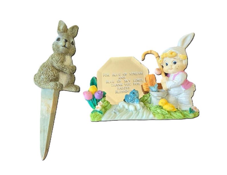 May include: A set of two Easter figurines. One is a brown rabbit with a white belly, the other is a girl in a white dress and pink jacket holding a fishing rod. The girl is standing on a base with a sign that reads "For the blue of stream and blue of sky Lord thank you for Easter blessing".
