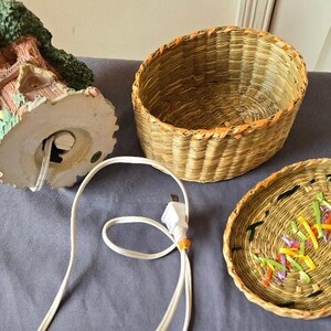 May include: A ceramic treehouse lamp with a woven basket base and lid. The lamp has a cord and plug. The basket lid has a woven design with colorful accents.