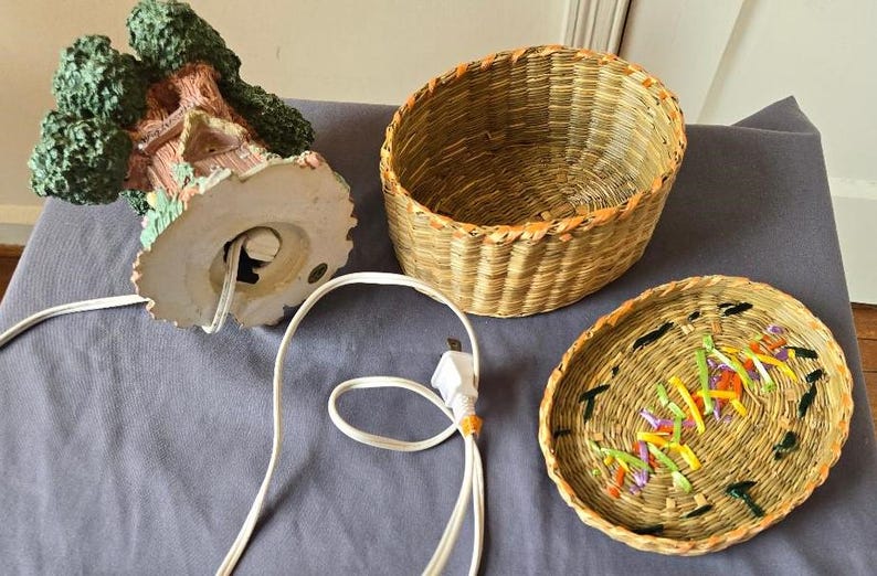 May include: A ceramic treehouse lamp with a woven basket base and lid. The lamp has a cord and plug. The basket lid has a woven design with colorful accents.