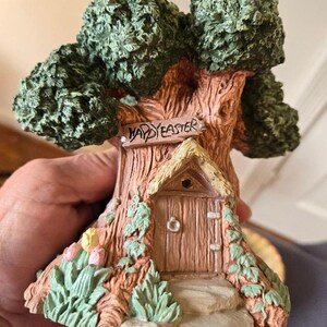 May include: A ceramic figurine of a tree house with a sign that reads "Happy Easter". The tree house has a small door and a thatched roof. The base of the figurine is decorated with green leaves and pink flowers.
