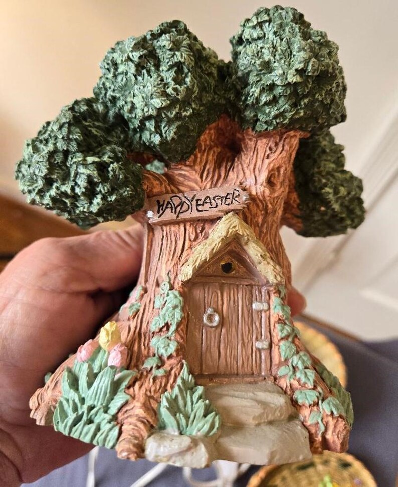 May include: A ceramic figurine of a tree house with a sign that reads "Happy Easter". The tree house has a small door and a thatched roof. The base of the figurine is decorated with green leaves and pink flowers.