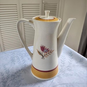 May include: A white ceramic coffee pot with a yellow and red floral design. The pot has a lid and a spout.