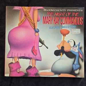 May include: A pink cartoon woman with a gun and a plunger holding a pink purse. The woman is wearing a pink dress and boots. The text on the image reads "Bloom County Presents The Night of the Mary Kay Commandos Featuring Smell-O-Toons".