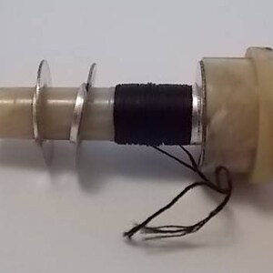 May include: A close-up of a white and black vintage sewing machine bobbin winder with a black thread spool and a white plastic base.