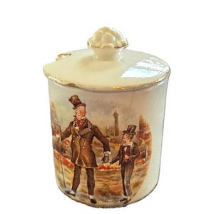 May include: A white ceramic jar with a lid and a decorative knob. The jar features a painted scene of two figures in Victorian-era attire, with a cityscape in the background. The rim and knob are accented with gold.