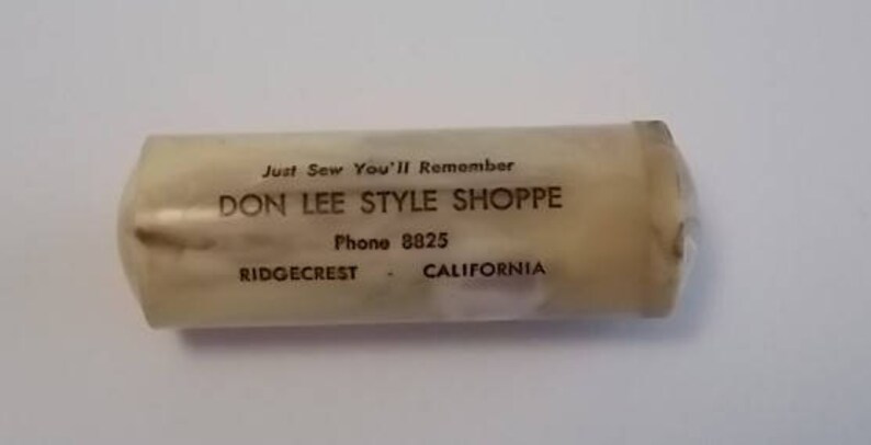 May include: A white plastic tube with the text "Just Sew You'll Remember Don Lee Style Shoppe Phone 8825 Ridgecrest - California" printed on it.