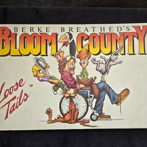 May include: A comic book cover featuring the title "Berke Breathed's Bloom County" with the subtitle "Loose Tails". The cover features a colorful illustration of characters from the comic strip, including a man in a wheelchair, a cat, a bird, and a rabbit.
