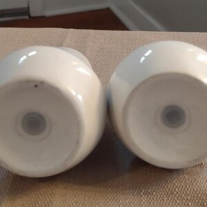 May include: Two white ceramic salt and pepper shakers with a round shape and a small hole in the bottom.