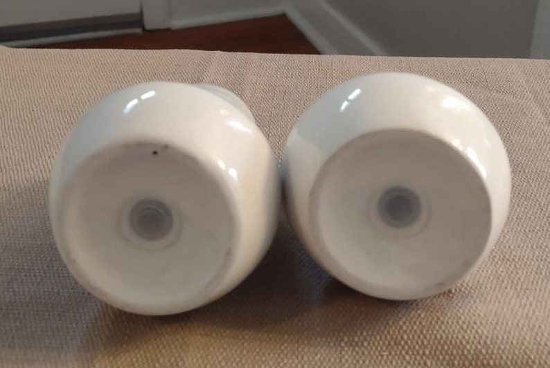 May include: Two white ceramic salt and pepper shakers with a round shape and a small hole in the bottom.