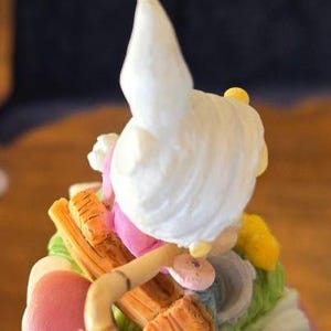 May include: A white ceramic figurine of a woman with a white bonnet and pink dress sitting on a wooden sled. The sled is decorated with brown and yellow paint. The figurine is surrounded by green and yellow fabric.