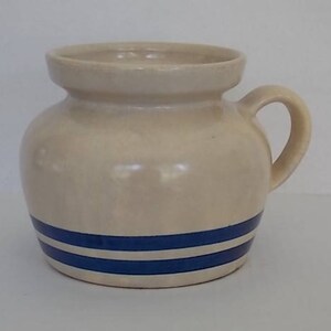 May include: A cream-colored ceramic pitcher with a blue and white striped design. The pitcher has a rounded body and a single handle.