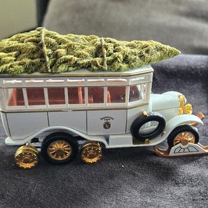 May include: White toy bus with a green Christmas tree on top. The bus has gold wheels and a black tire on the back. The bus has a sign that says "Bellissima Line" on the side.