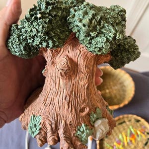 May include: A ceramic tree sculpture with a brown trunk and green leaves. The base of the tree is decorated with green leaves and a small gray rock.