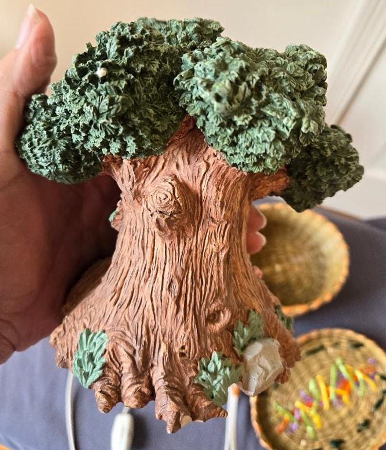 May include: A ceramic tree sculpture with a brown trunk and green leaves. The base of the tree is decorated with green leaves and a small gray rock.