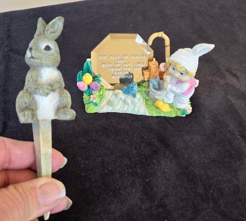 May include: A gray rabbit figurine with a white belly and a blue and white figurine of a girl holding a bucket of fish. The background is a blue and gold plaque with the text "For Blue of Stream and Blue of Sky I Give Thank You For Blessings".