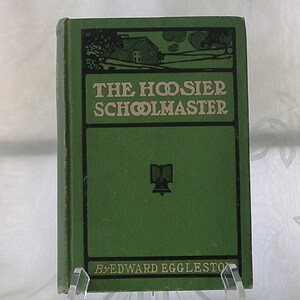 May include: A green hardcover book with the title "The Hoosier Schoolmaster" by Edward Eggleston. The cover features a black and white illustration of a house and a book with a bell.