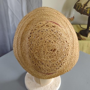 Puede incluir: Un sombrero de paja con un dise&ntilde;o tejido y un borde rojo alrededor del borde. El sombrero est&aacute; en un soporte de exhibici&oacute;n blanco.