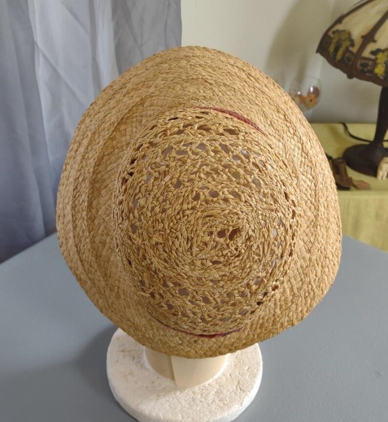 Puede incluir: Un sombrero de paja con un dise&ntilde;o tejido y un borde rojo alrededor del borde. El sombrero est&aacute; en un soporte de exhibici&oacute;n blanco.