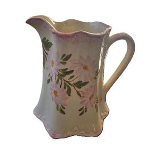 May include: A white ceramic pitcher with a pink rim and floral design. The pitcher has a scalloped edge and a handle.