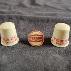 May include: Three white plastic thimbles with the Texaco logo printed in red. The thimbles are on a gray fabric surface.