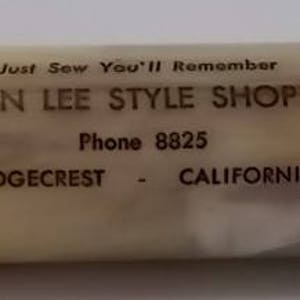 May include: A white plastic cylinder with brown text that reads "Just Sew You'll Remember Don Lee Style Shoppe Phone 8825 Ridgecrest - California".