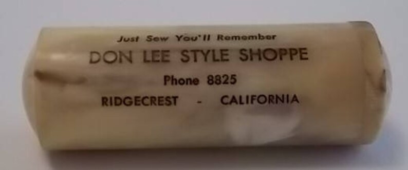 May include: A white plastic cylinder with brown text that reads "Just Sew You'll Remember Don Lee Style Shoppe Phone 8825 Ridgecrest - California".
