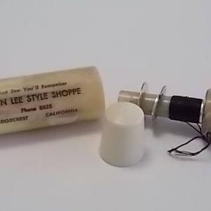 May include: A white plastic container with the text "Don Lee Style Shoppe" and the phone number "6223" printed on it. The container is open and a white plastic spool with a black thread is visible. The spool is attached to a white plastic cap.