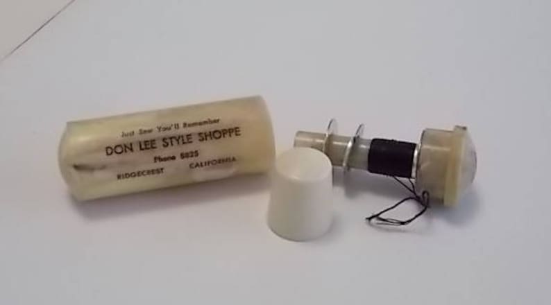 May include: A white plastic container with the text "Don Lee Style Shoppe" and the phone number "6223" printed on it. The container is open and a white plastic spool with a black thread is visible. The spool is attached to a white plastic cap.