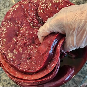May include: A close-up of a large, raw, red liver being held in a gloved hand. The liver is in a bowl and appears to be soaking in a red liquid.