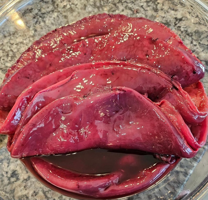 May include: A close-up of a bowl of raw, red beef liver. The liver is sliced and arranged in a bowl.