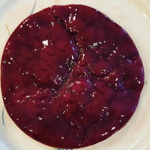 May include: A close-up of a round, red, and purple food item on a white plate. The food has a smooth, shiny surface and appears to be a type of meat.