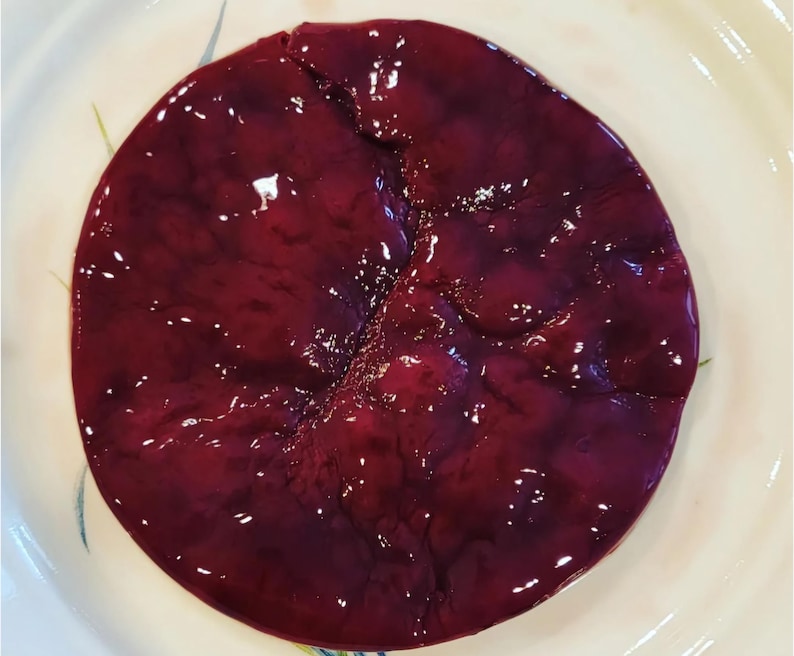 May include: A close-up of a round, red, and purple food item on a white plate. The food has a smooth, shiny surface and appears to be a type of meat.