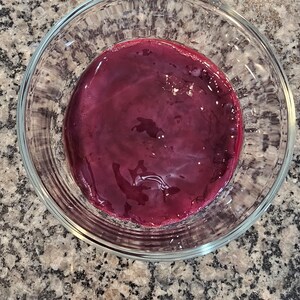 May include: A glass bowl filled with a deep red, almost purple, liquid. The liquid is smooth and glossy, reflecting light.