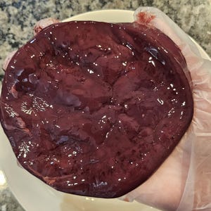 May include: A close-up of a raw, dark red beef liver. The liver is smooth and shiny, with a slightly textured surface.