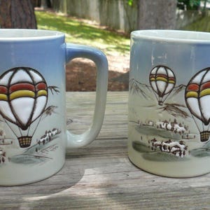 May include: Two ceramic mugs with a light blue gradient. Each mug features a detailed illustration of hot air balloons with white, yellow, and red accents, floating above a landscape with mountains and houses. The mugs have large handles.