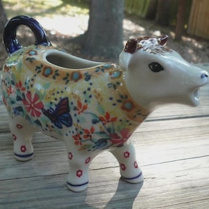 Pode incluir: Um cremeira de vaca de cerâmica com um padrão floral e borboleta. A cremeira tem uma cabeça e pernas brancas, com uma alça azul escura e um bico para servir. O corpo é decorado com flores vermelhas, azuis e amarelas.