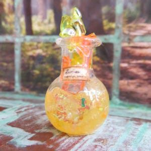 May include: A small, clear glass bottle with a cork stopper and a yellow liquid inside. The bottle is decorated with iridescent glitter and a small, orange, plastic flower. The label on the bottle reads "Surreal".