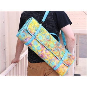 K&ouml;nnte beinhalten: Eine Yogamatten-Tasche mit floralem Muster und hellblauem Gurt und Besatz. Die Tasche wird &uuml;ber der Schulter getragen und weist eine lebendige Mischung aus Gelb-, Blau- und Lilat&ouml;nen auf. Sie ist f&uuml;r eine gerollte Yogamatte konzipiert.