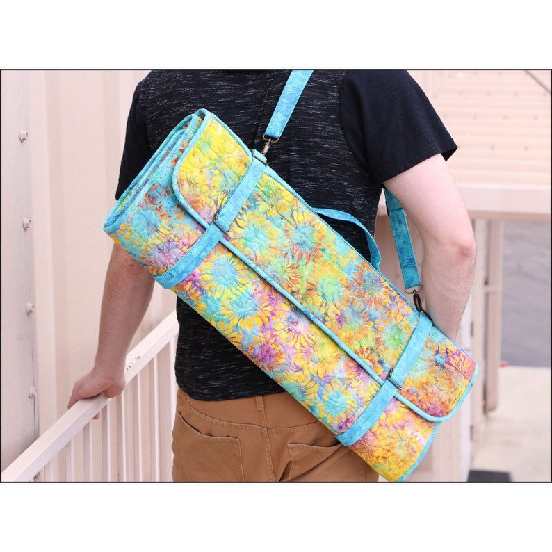 K&ouml;nnte beinhalten: Eine Yogamatten-Tasche mit floralem Muster und hellblauem Gurt und Besatz. Die Tasche wird &uuml;ber der Schulter getragen und weist eine lebendige Mischung aus Gelb-, Blau- und Lilat&ouml;nen auf. Sie ist f&uuml;r eine gerollte Yogamatte konzipiert.