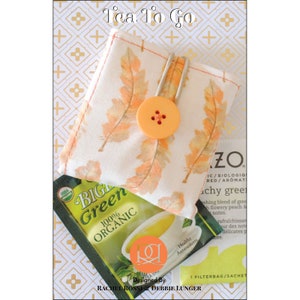 May include: A fabric tea bag holder with a feather pattern in orange and peach, secured with a button. The words "Tea To Go" are at the top. A green tea bag is visible below.