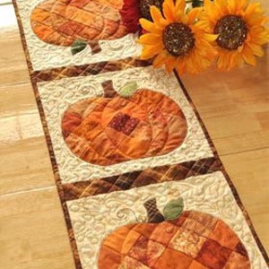 K&ouml;nnte beinhalten: Gesteppter Tischl&auml;ufer mit vier orangefarbenen K&uuml;rbismotiven und gr&uuml;nen Stielen. Die K&uuml;rbisse sind vor einem cremefarbenen Hintergrund mit floralem Muster angeordnet. Eine rote Blume und zwei Sonnenblumen sind an der Seite platziert.