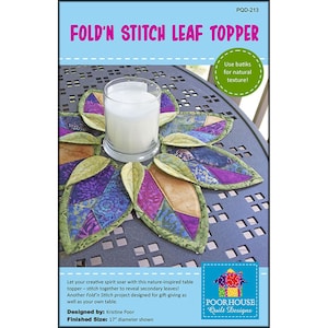 May include: A handmade leaf-shaped table topper with a candle holder in the center. The topper is made of colorful fabric pieces, including purple, green, and gold. The text "FOLD'N STITCH LEAF TOPPER" is displayed at the top. The finished size is 17 inches in diameter.