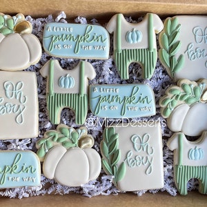 Op de afbeelding: Een set van 10 gedecoreerde suikerkoekjes in een doos. De koekjes zijn versierd met groene bladeren, pompoenen en de tekst "Oh boy" en "A little pumpkin is on the way".