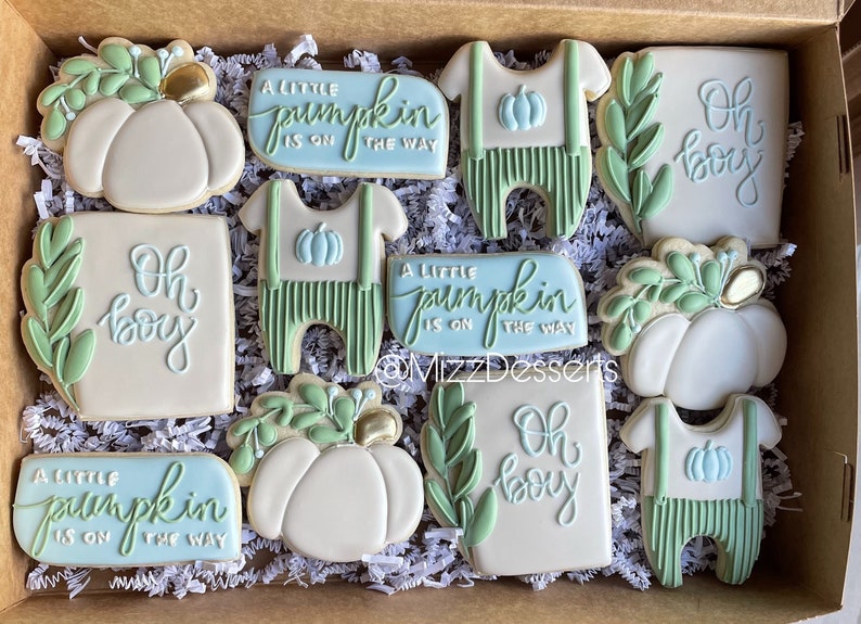 Op de afbeelding: Een set van 10 gedecoreerde suikerkoekjes in een doos. De koekjes zijn versierd met groene bladeren, pompoenen en de tekst "Oh boy" en "A little pumpkin is on the way".