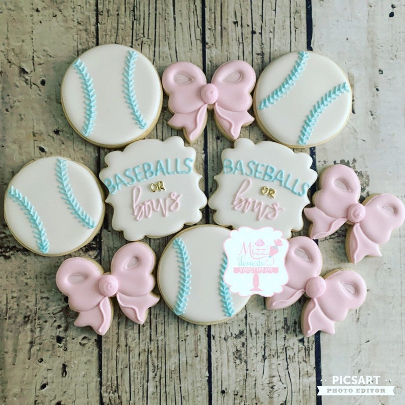 K&ouml;nnte beinhalten: Ein Set aus neun dekorierten Zuckerkeksen. Vier Kekse sind in Form von Baseballs mit blauen N&auml;hten und wei&szlig;em Hintergrund. Zwei Kekse sind quadratisch mit dem Text "BASEBALLS OR BOWS" in blauer und goldener Schrift. Drei Kekse sind in Form von rosa Schleifen mit wei&szlig;em Zentrum.
