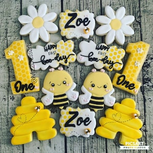 May include: A set of 10 decorated sugar cookies featuring a bee theme. The cookies are yellow and white with black accents. The cookies feature the words "Zoe", "One", and "Happy First Bee-day".