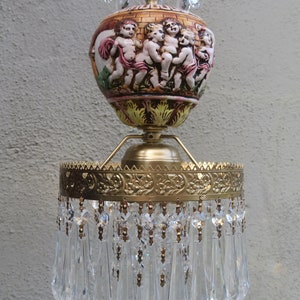 May include: A vintage chandelier with a ceramic center featuring cherubs. The fixture has a gold-toned metal frame and is adorned with clear crystal pendants and pink crystal accents. The design is ornate and decorative.