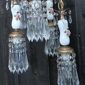 May include: A multi-light chandelier featuring white porcelain bodies adorned with small pink rose accents. Each light fixture is suspended by a gold-toned metal chain and decorated with numerous clear glass crystals and hanging teardrop prisms.
